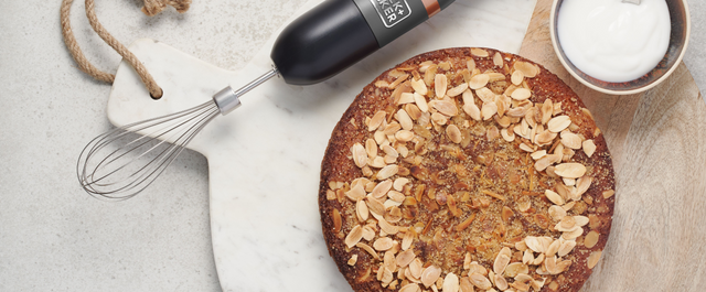 Grey, BLACK+DECKER kitchen wand with whisk attachment lying next to a marble plate with a gluten free cherry and almond cake