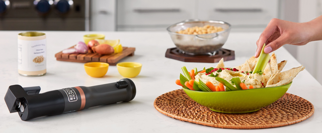 Zoomed in shot of talent dipping celery into prepared hummus, featuring the grey, BLACK+DECKER kitchen wand can opener