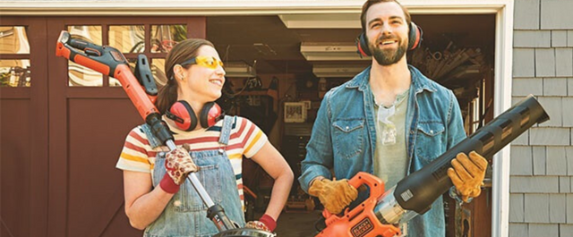 Woman and man standing in front of garage while holding outdoor power tools, such as a string trimmer and leaf blower