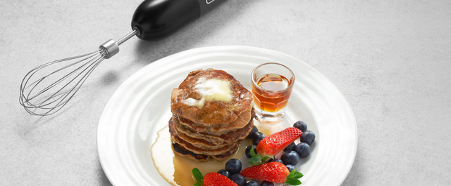 Grey, BLACK+DECKER kitchen wand with whisk attachment lying next to a plate of pancakes, mixed berries, and maple syrup