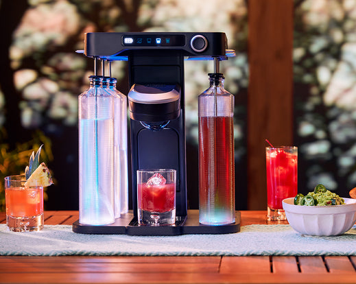 blue lights illuminated on the bev by BLACK+DECKER™ cordless cocktail maker sitting on a wooden patio table with drinks and appetizers
